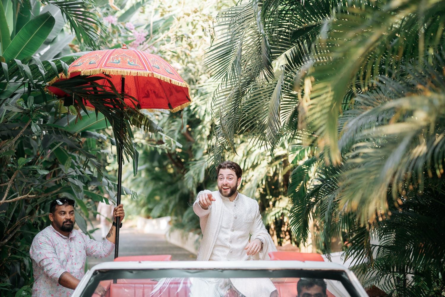 Groom in white sherwani stands through sunroof of car under red umbrella during Indian wedding baraat ceremony.