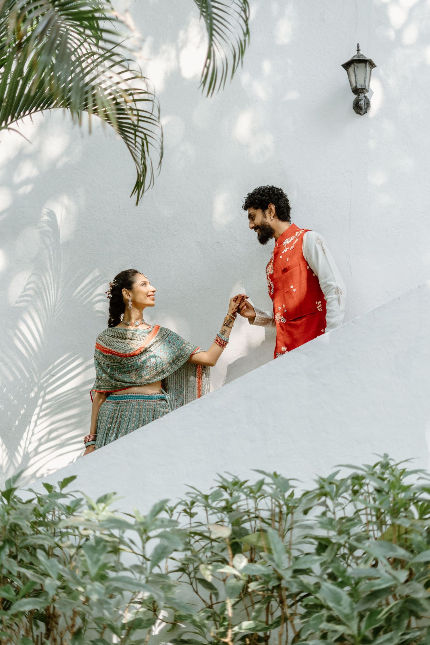 Indian couple in traditional attire holding hands on a sunlit terrace surrounded by lush tropical greenery.