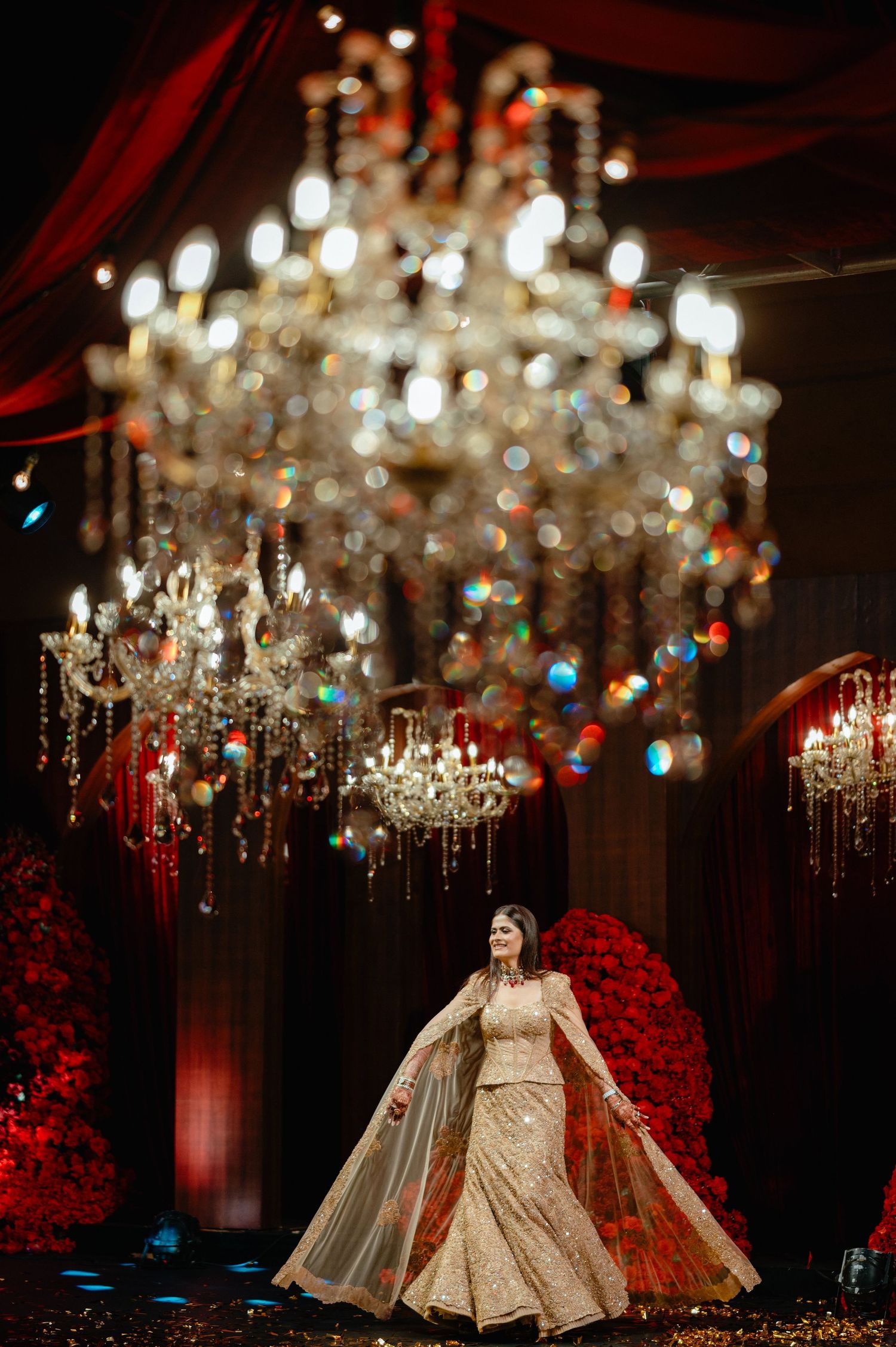 Bride in gold lehenga stands under grand crystal chandeliers at an opulent Indian wedding reception with red floral decor.