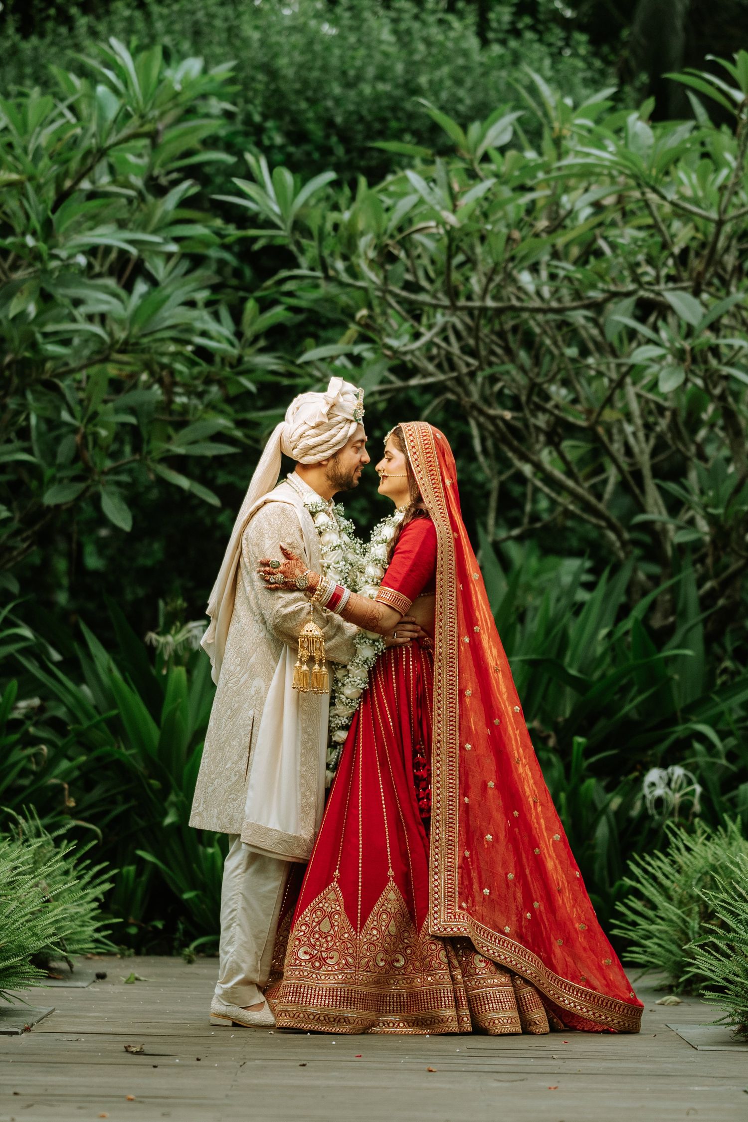 Indian bride in red lehenga and groom in cream sherwani share a romantic moment in a lush garden setting.