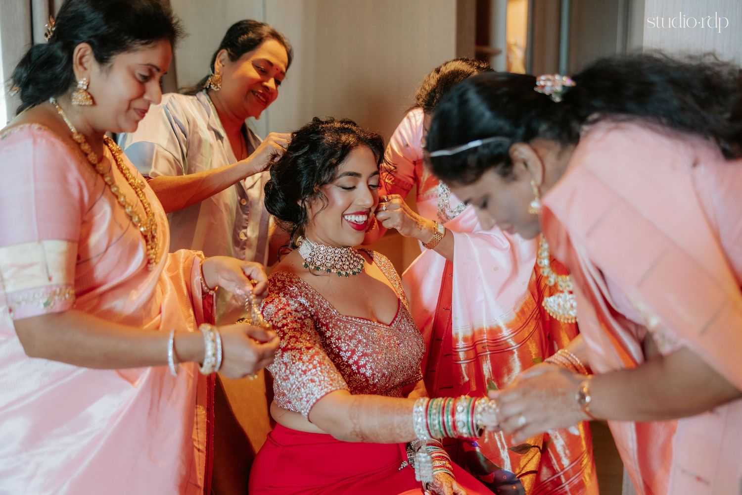 Bride in red lehenga surrounded by women in pink sarees during a traditional Indian wedding getting ready.