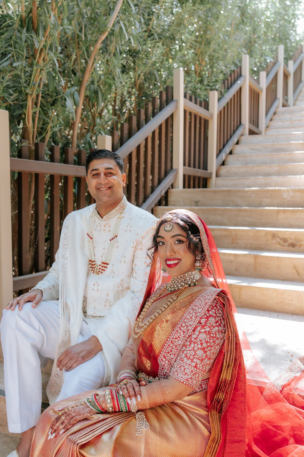 Indian bride in red saree and groom in white sherwani pose together on outdoor staircase at traditional Hindu wedding.