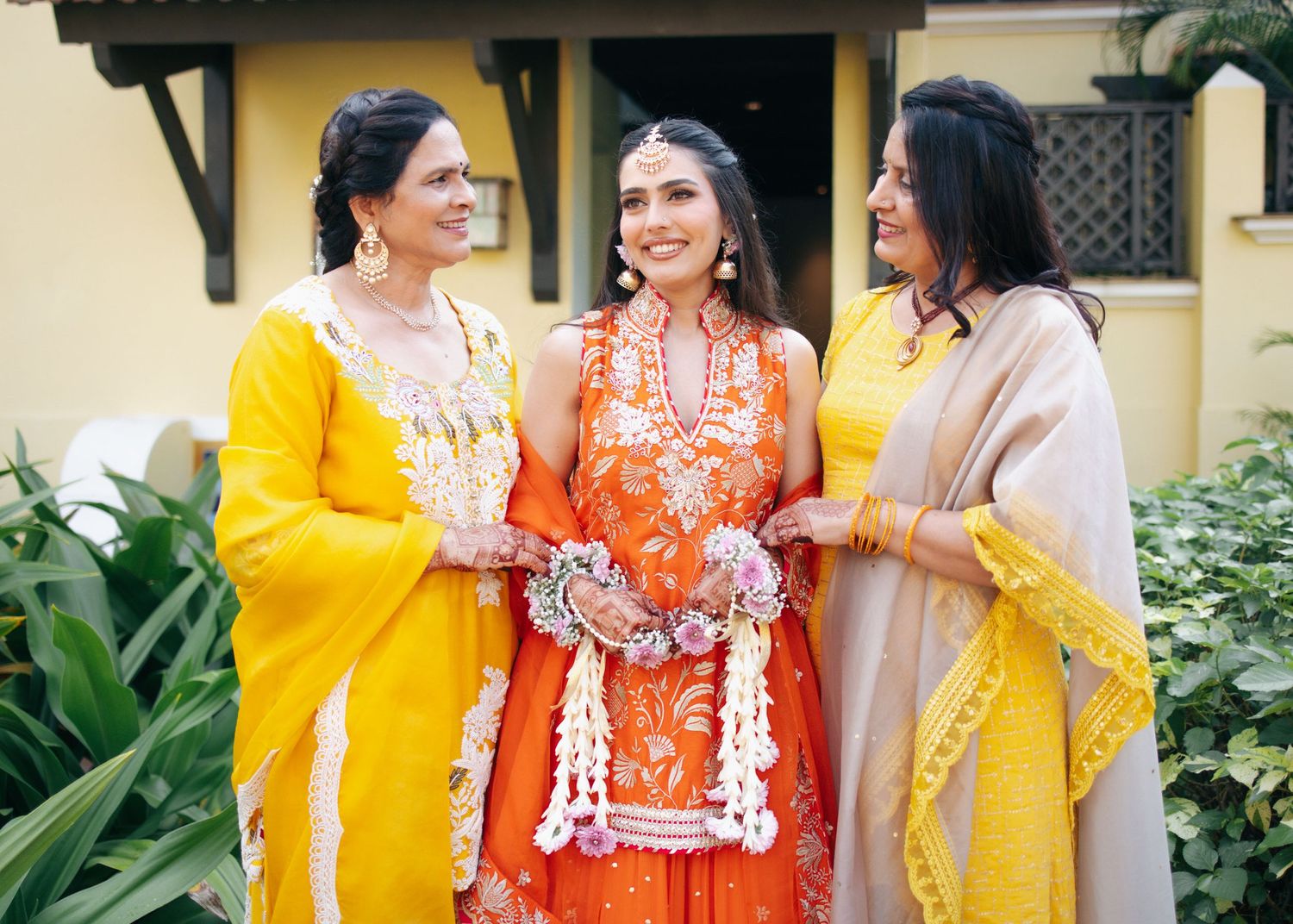 Three people in traditional South Asian attire including yellow and orange saris pose together in a garden setting.