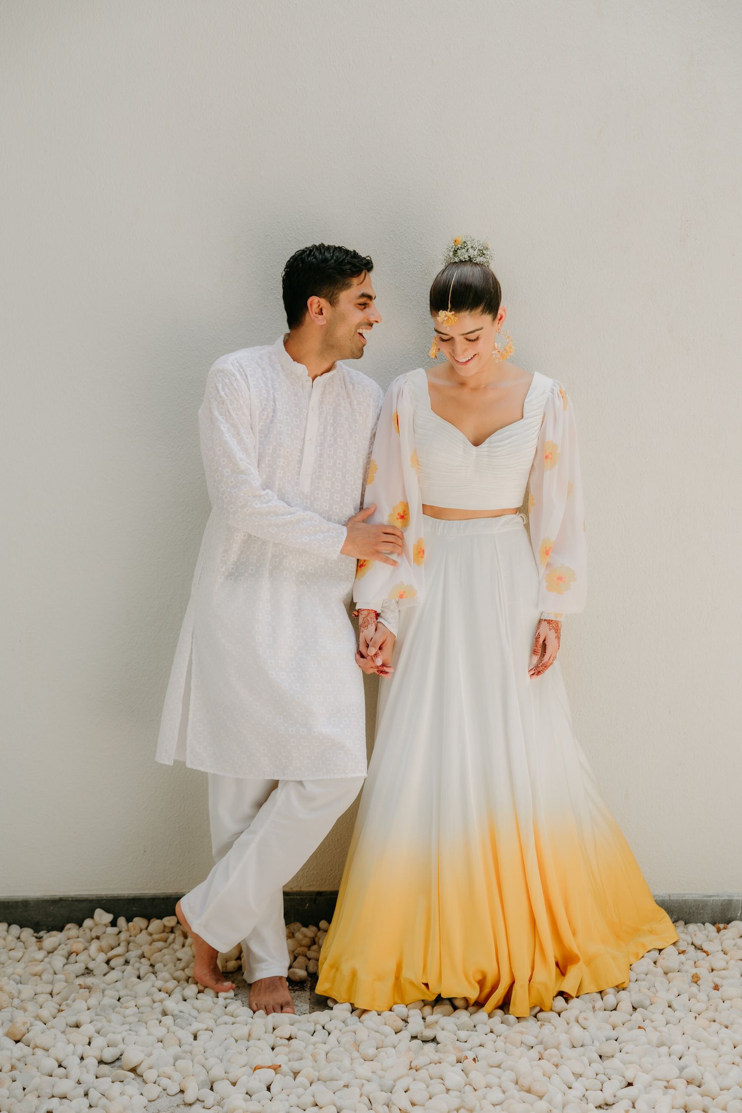 Couple in white attire poses against wall, bride wearing unique ombre yellow and white gown.