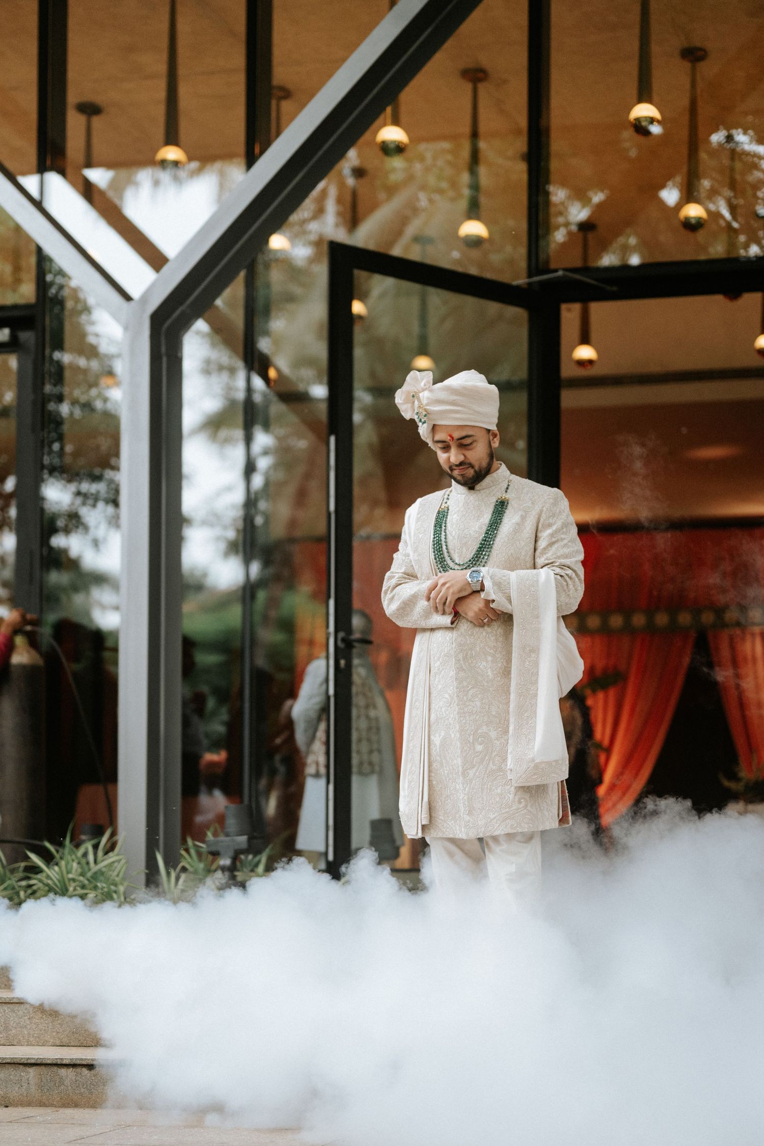 Indian groom in white sherwani and turban checks watch amid dramatic fog at elegant wedding venue.