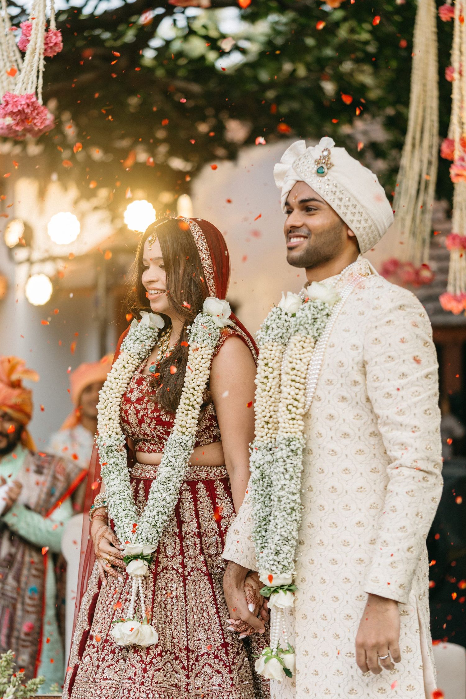 Indian bride in red lehenga and groom in white sherwani exchange garlands at traditional Hindu wedding ceremony.