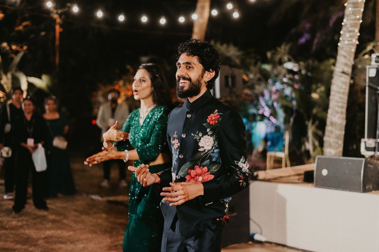 Couple in elegant Indian attire at an outdoor evening wedding celebration with string lights.