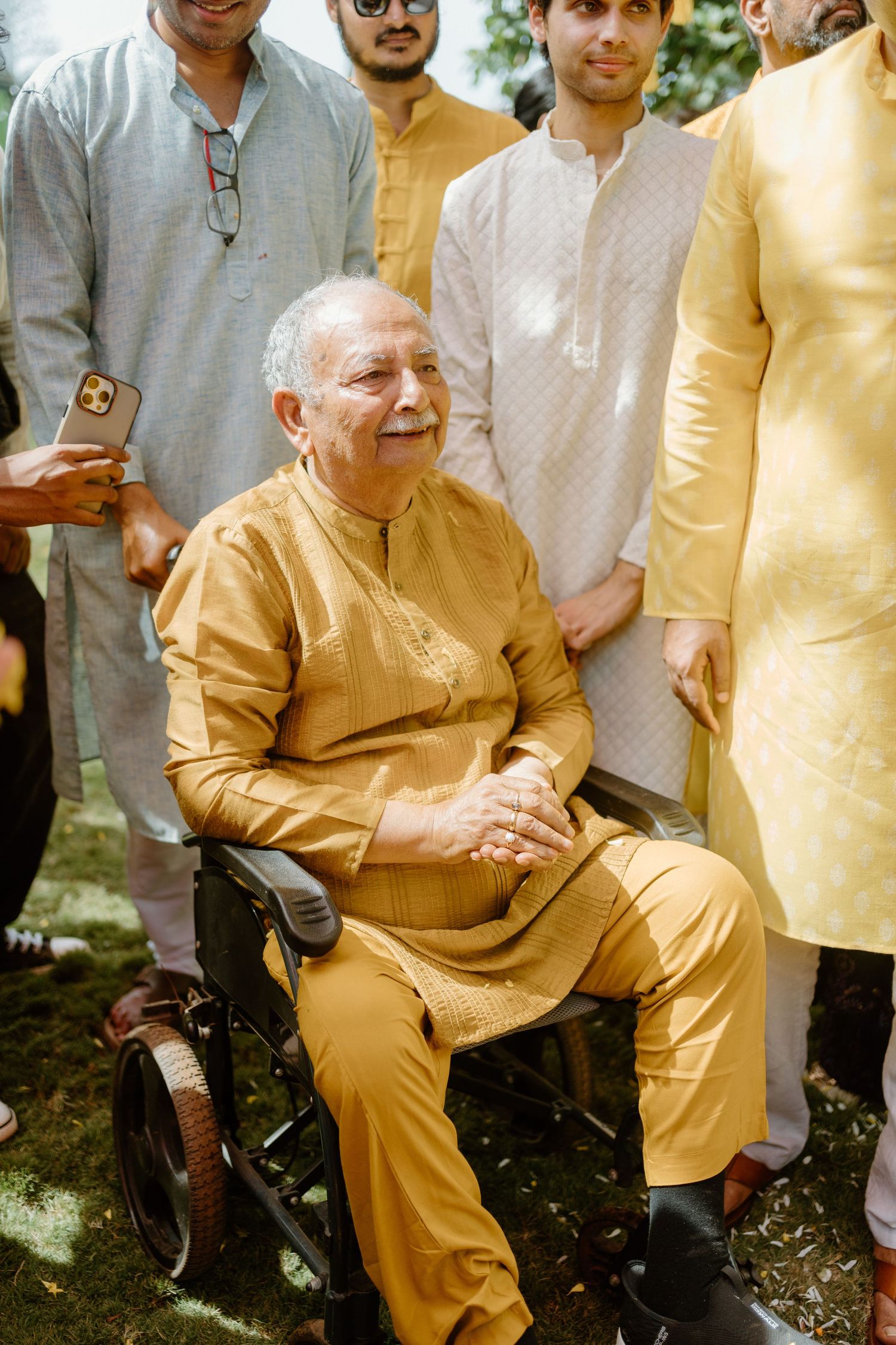Elderly man in golden kurta sitting in wheelchair surrounded by family at Indian celebration.