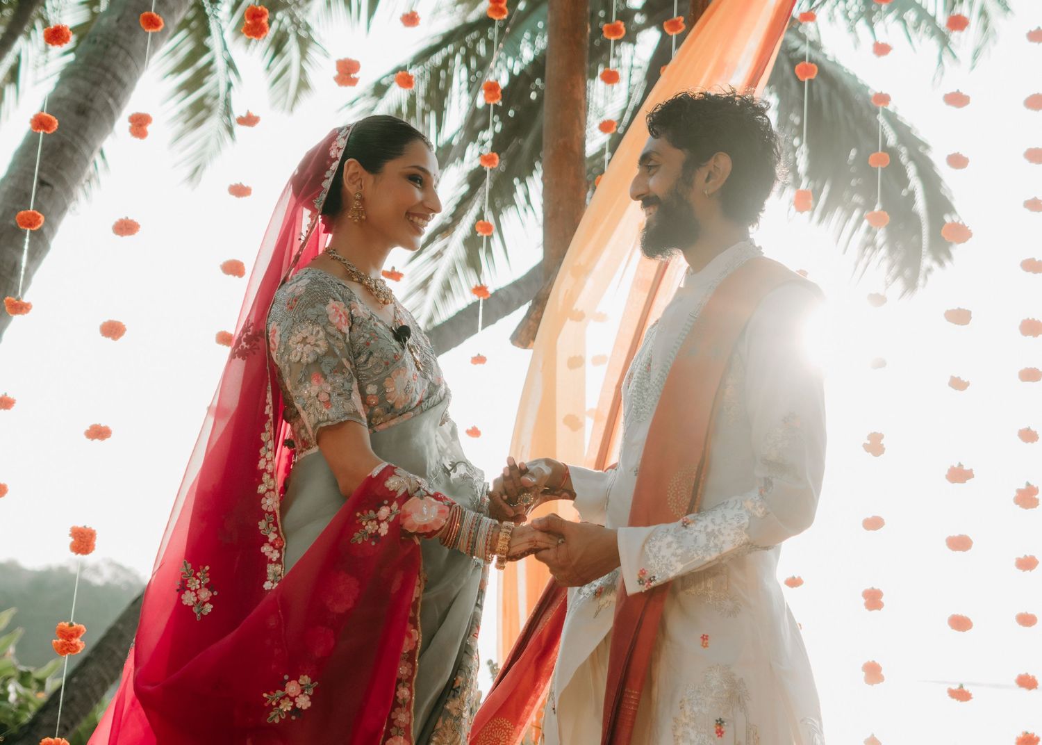 Couple in traditional Indian wedding attire holding hands under floral decorations with warm sunlight.
