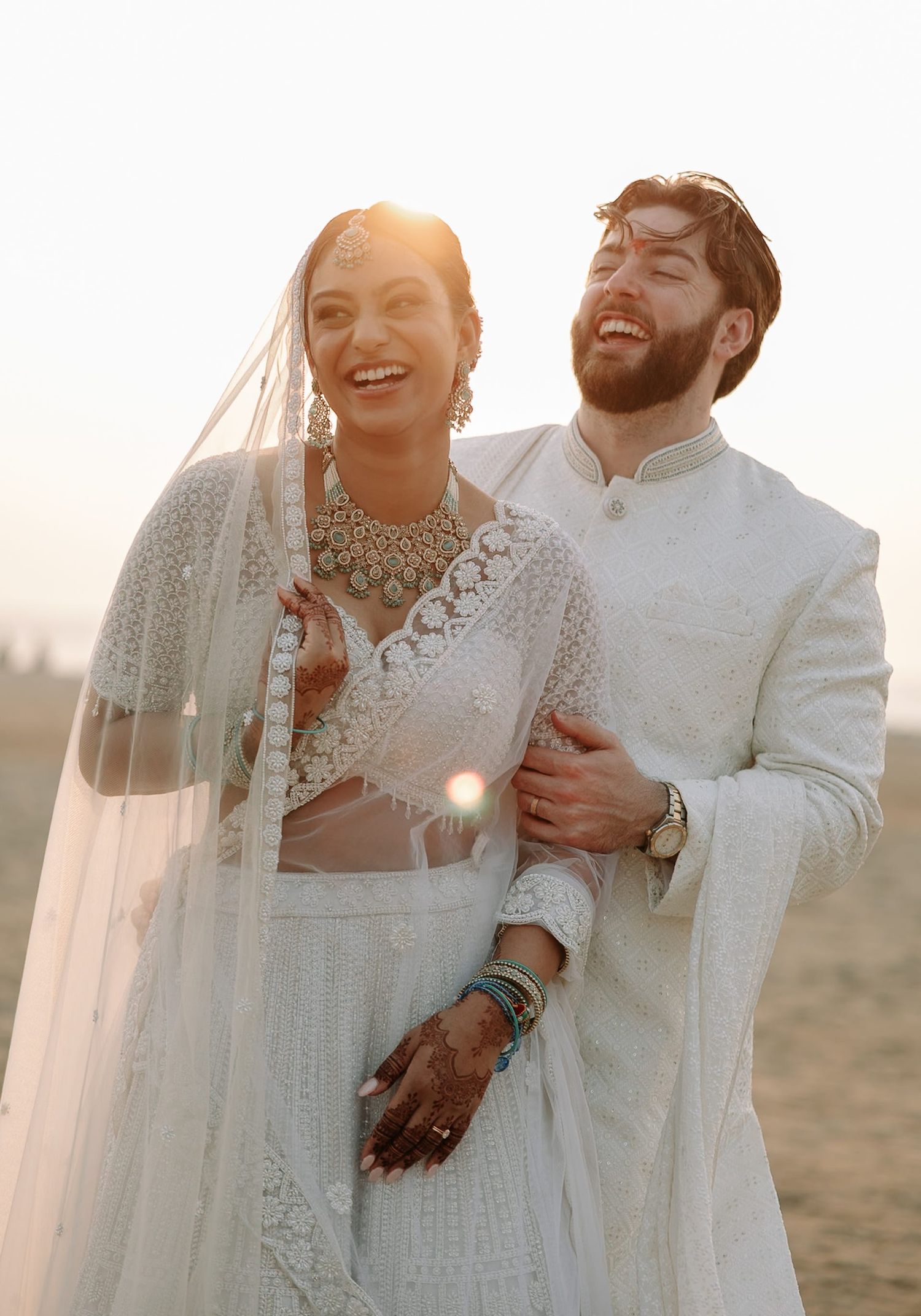Joyful couple in white Indian wedding attire, bride in lehenga with veil and groom in sherwani, golden sunset backdrop.