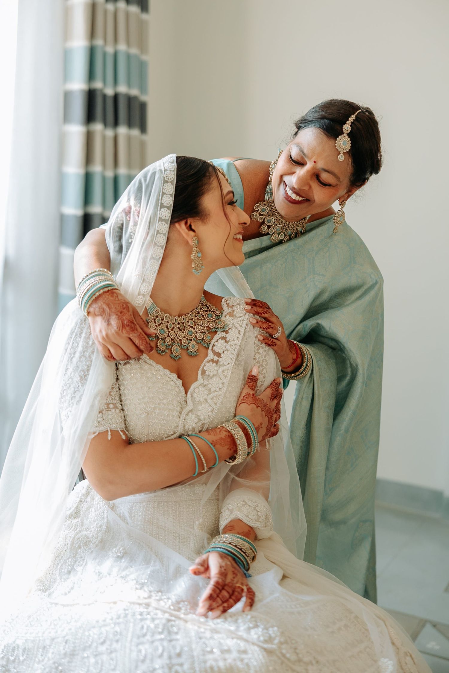 Two Indian women in traditional bridal attire share a joyful embrace, adorned with jewelry and mehndi.