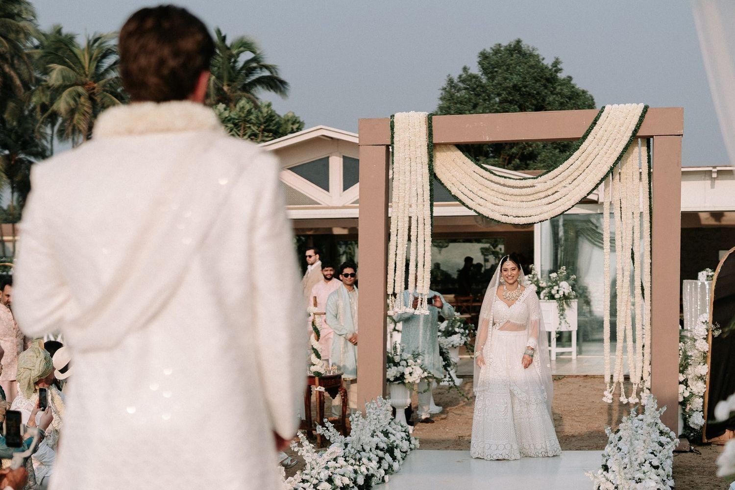 Bride walks down the aisle toward groom at an elegant outdoor wedding ceremony with floral arch decor.