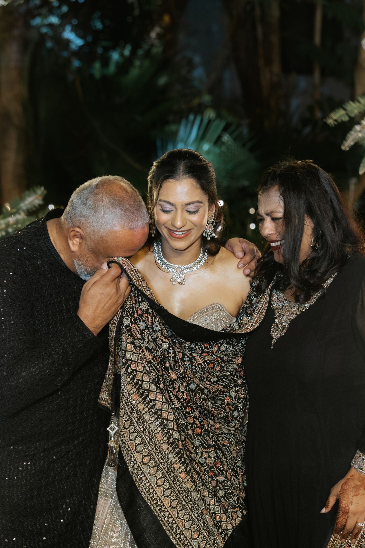 Bride in embroidered black lehenga with silver jewelry shares tender moment with parents at Indian wedding.