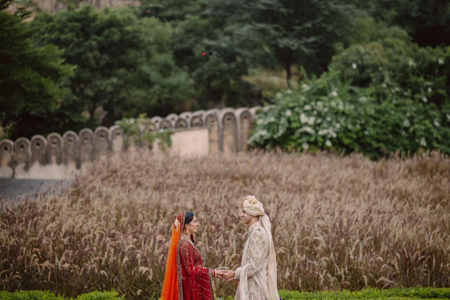 Indian bride in red lehenga and groom in ivory sherwani hold hands in a golden wheat field at a heritage venue.
