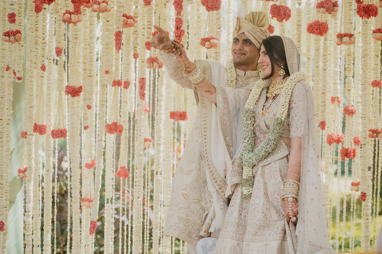 A couple in traditional white and mint wedding attire pose under a backdrop of hanging red floral decorations.