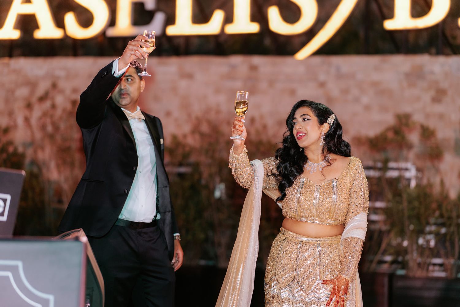 Indian bride in gold lehenga and guest raising champagne glasses in toast at outdoor wedding reception.