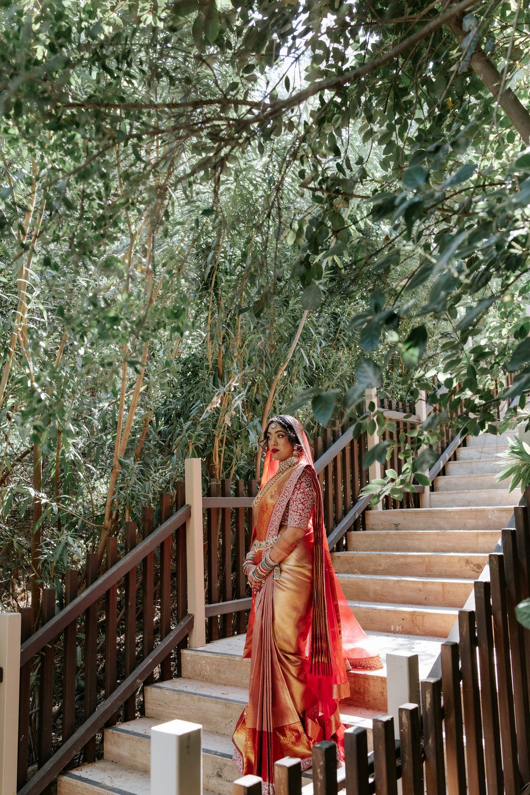 Elegant Indian bride in traditional red and gold saree posing on outdoor staircase with green trees.