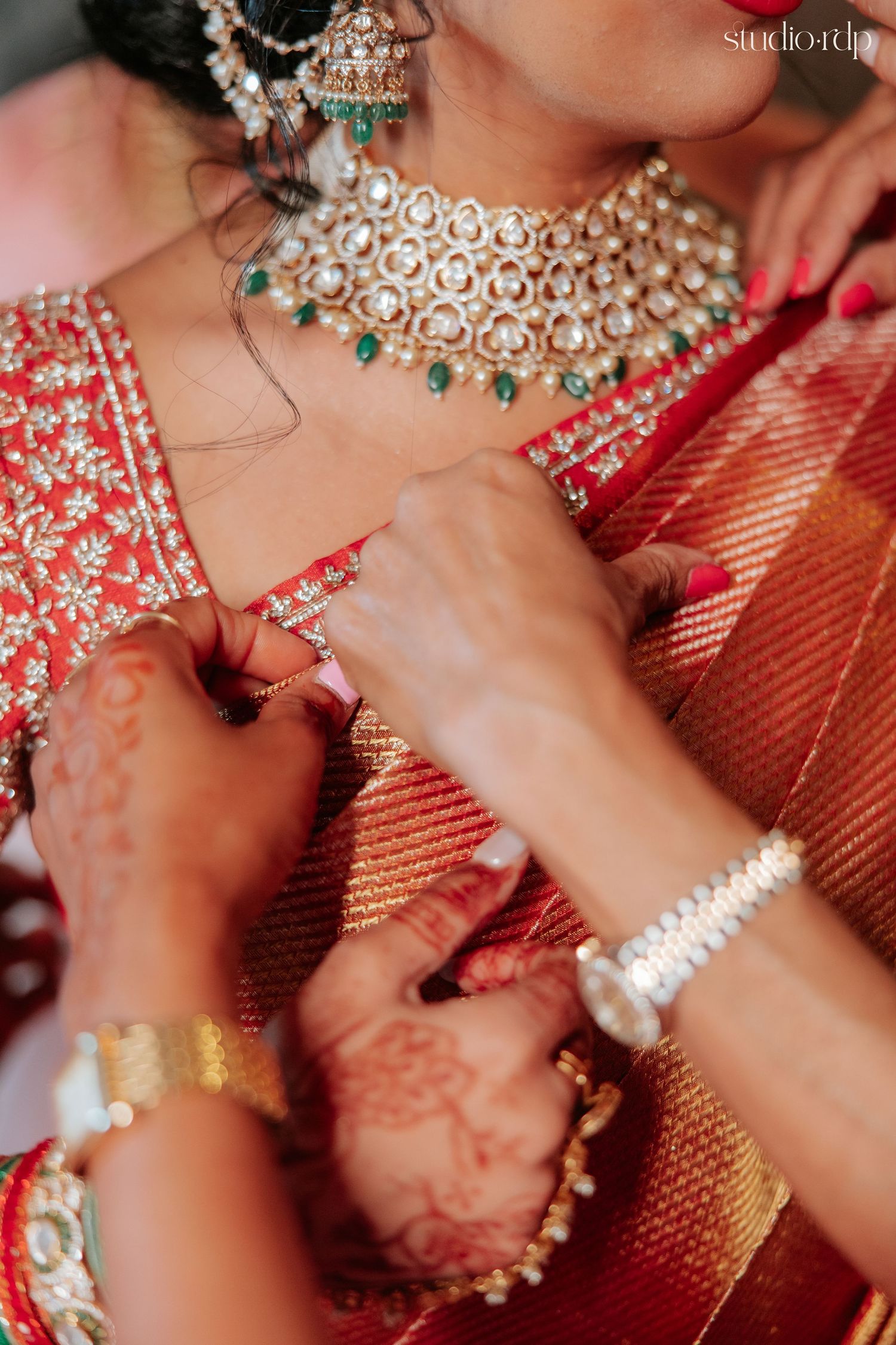 Indian bride in red saree with gold kundan necklace and mehndi hands being assisted by family during wedding preparation.