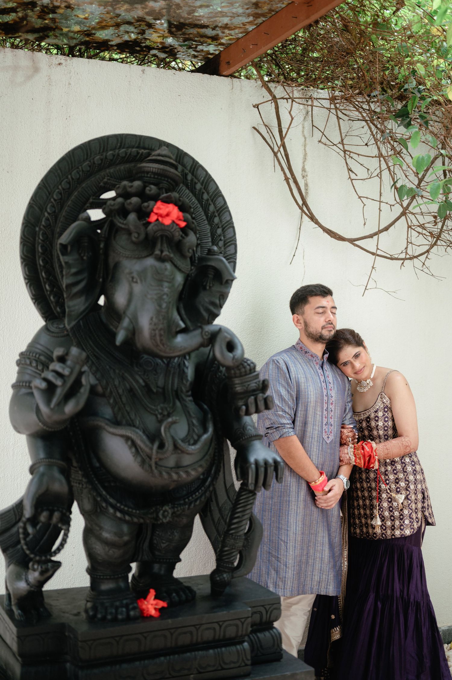 Couple in traditional Indian attire posing beside a large black Ganesha statue decorated with red flowers.