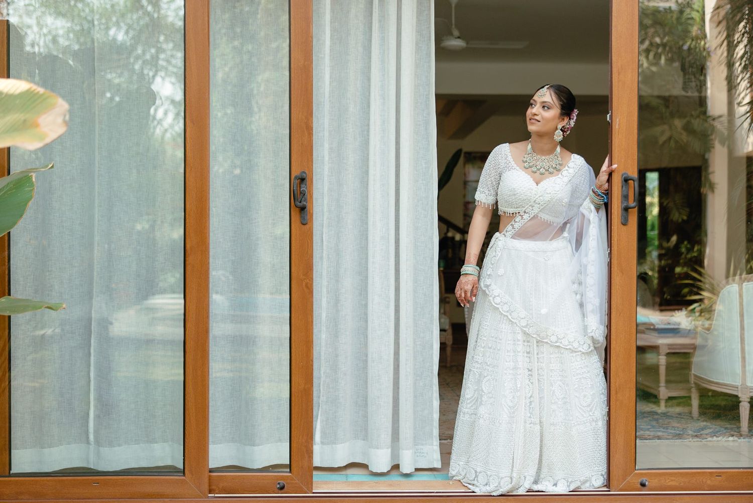 Bride in white lehenga choli with silver jewelry posing elegantly at wooden glass doors during wedding.