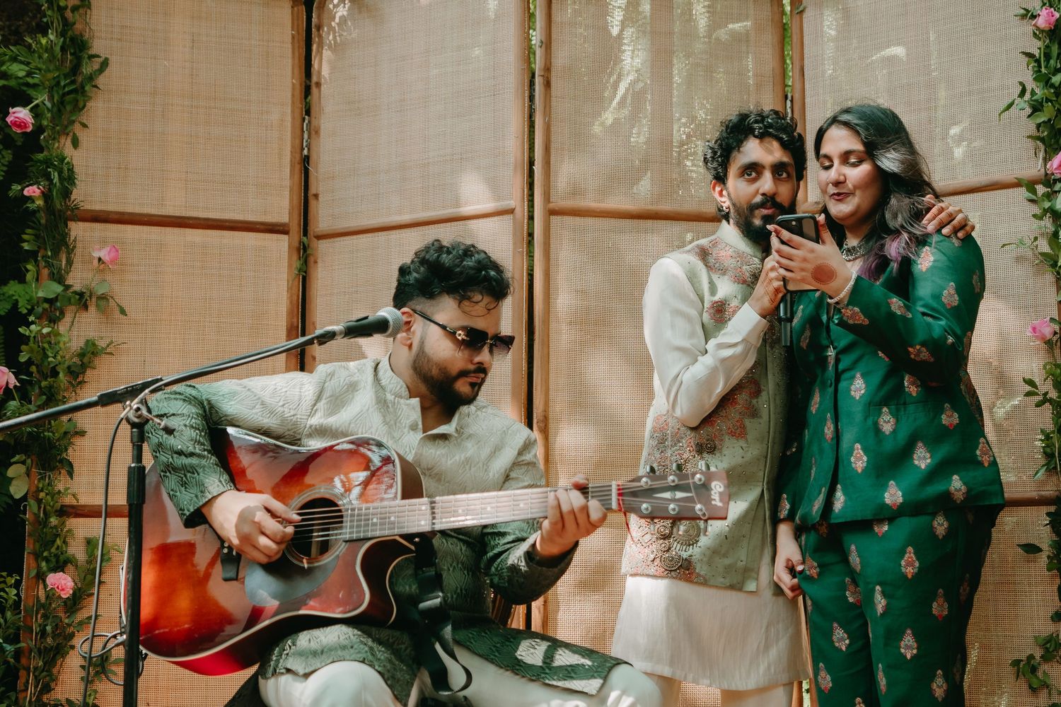 Musician playing acoustic guitar at Indian wedding celebration with two guests singing in traditional attire.