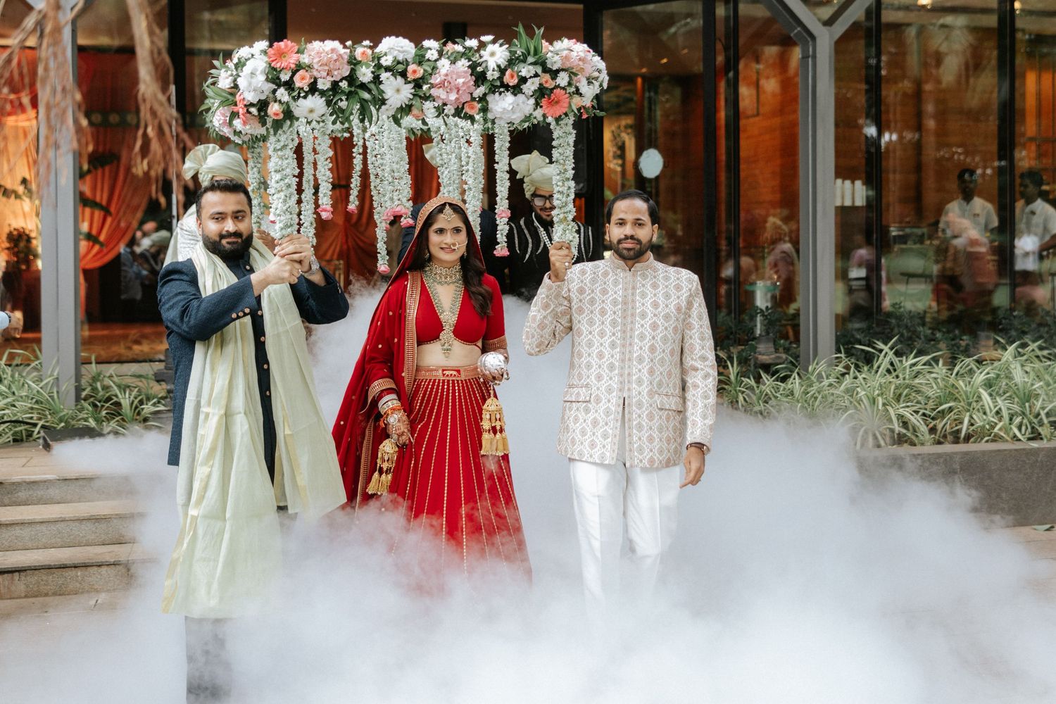 Bride in red lehenga with two grooms during Indian wedding entrance ceremony with floral decor and dramatic fog effect.