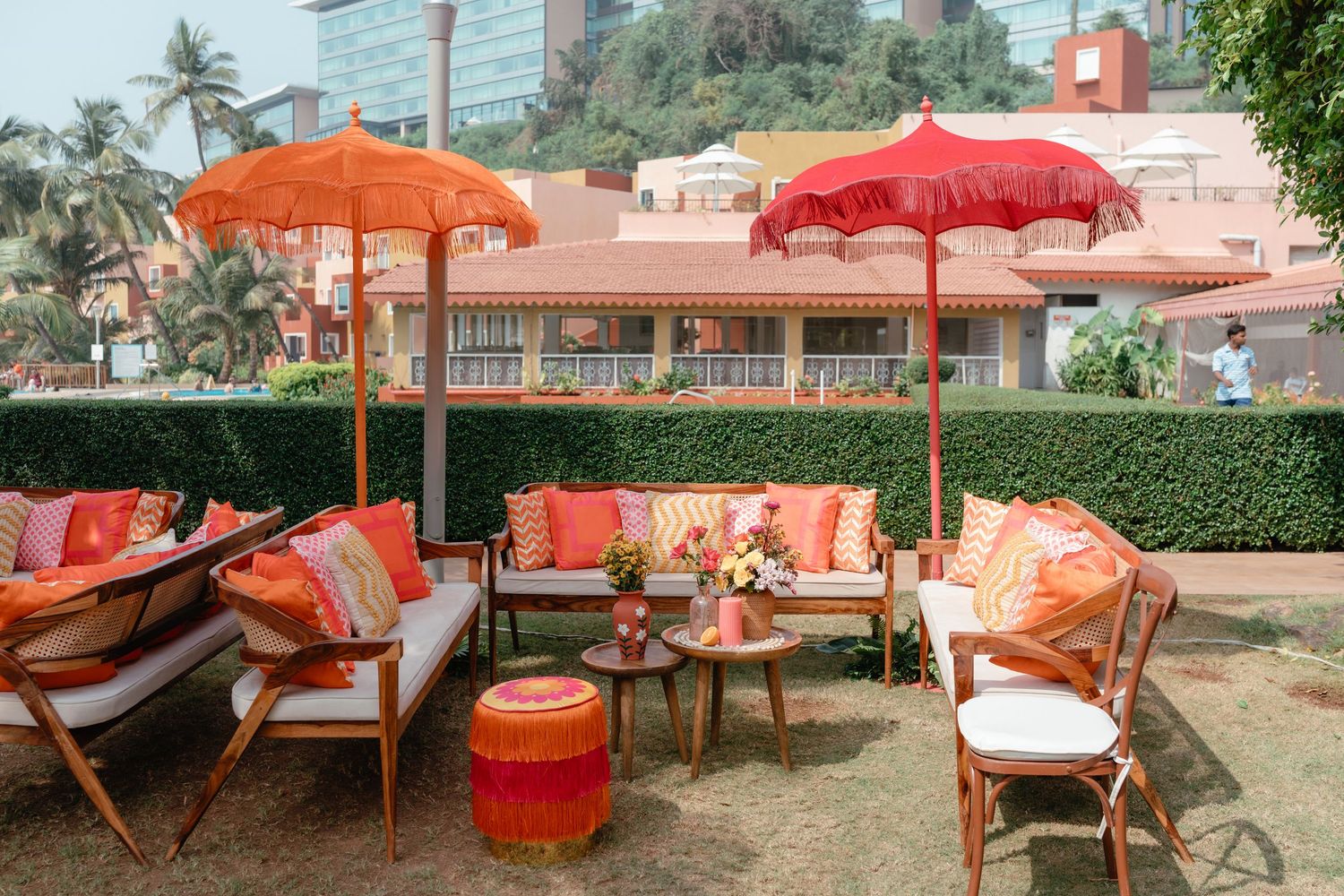 Tropical outdoor lounge area with orange and red umbrellas, cushioned seating, and palm trees in the background.