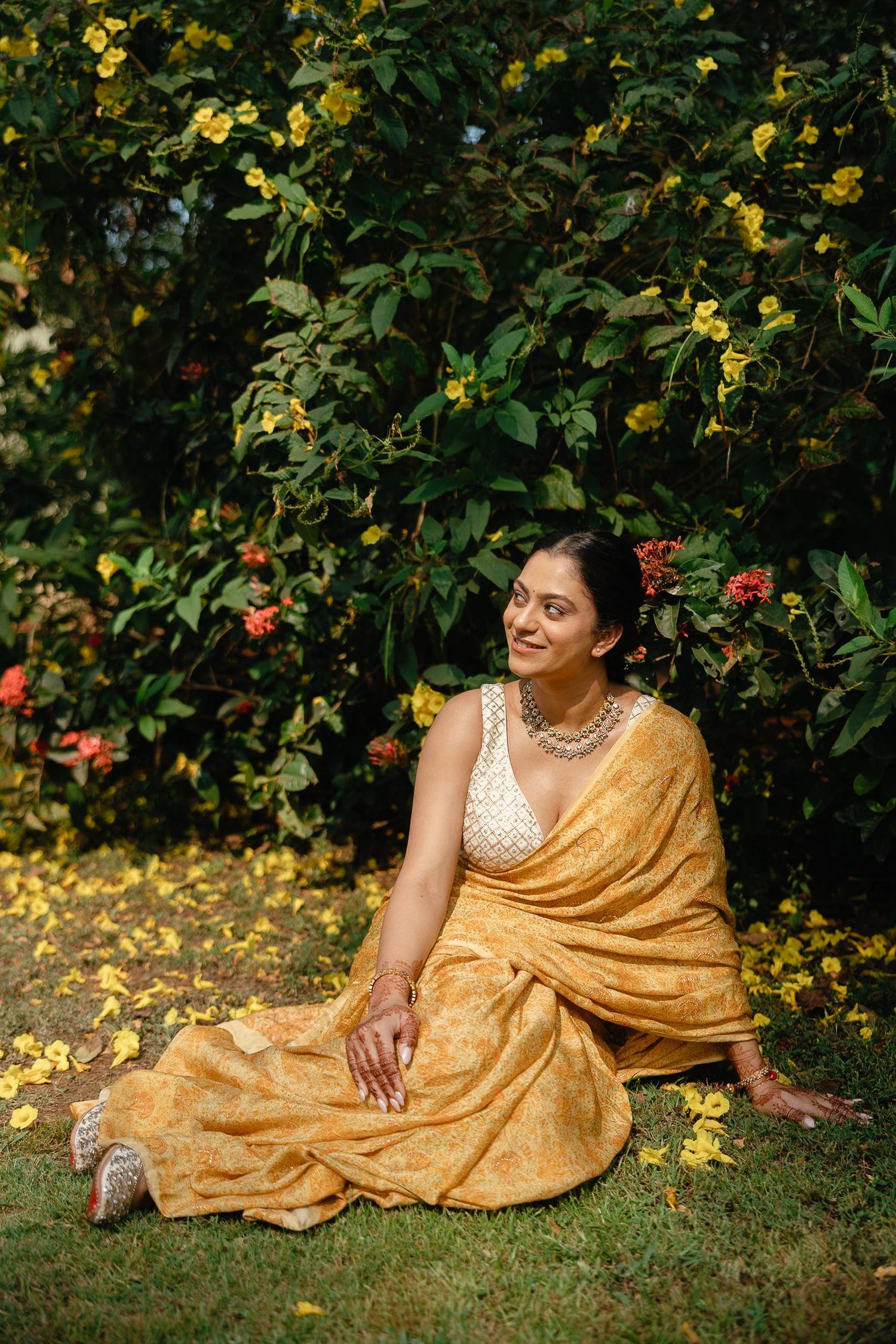 Woman in golden yellow saree sitting gracefully on grass beside blooming yellow flowers.