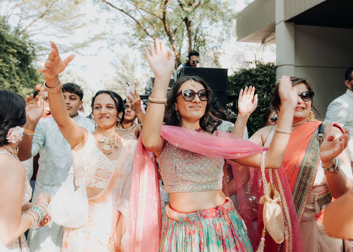 A festive wedding party wearing pastel dresses celebrates joyfully with raised hands during an outdoor celebration.
