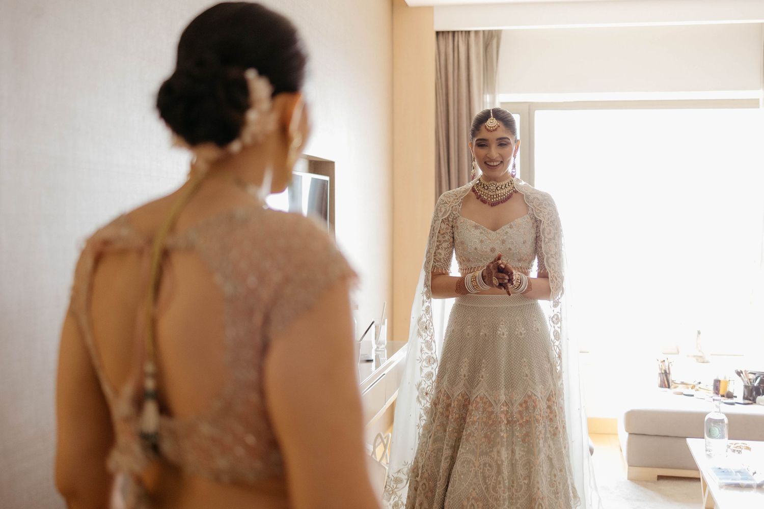 Bride wearing an elegant lace wedding gown looking in the mirror while getting ready in a bright bedroom during wedding morning preparations