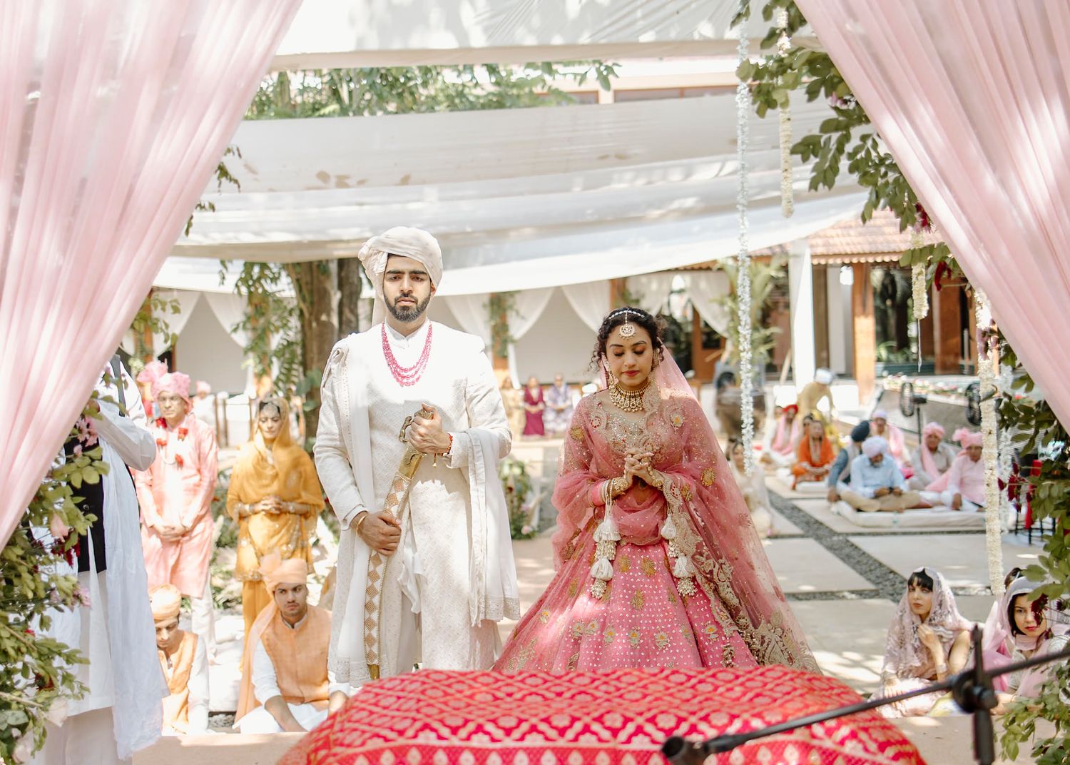 Bride in pink lehenga and groom in white sherwani walk together at an elegant outdoor Indian wedding ceremony.