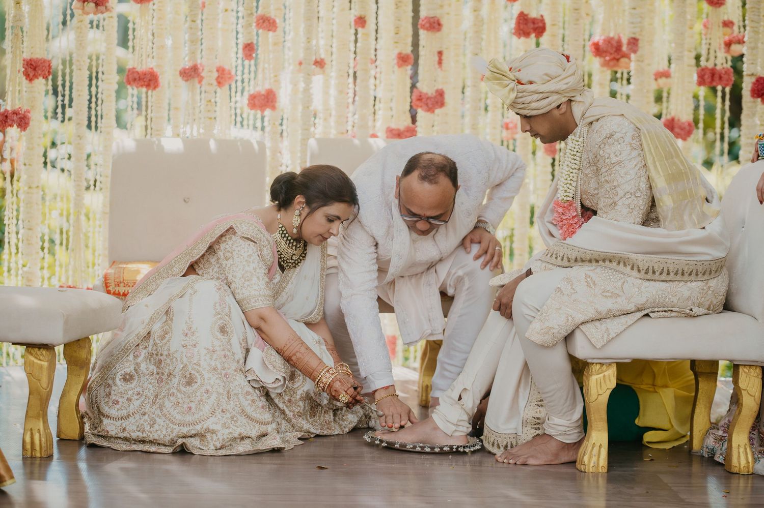 Traditional Indian wedding ceremony participants perform ritual under floral decorations in white and cream attire.