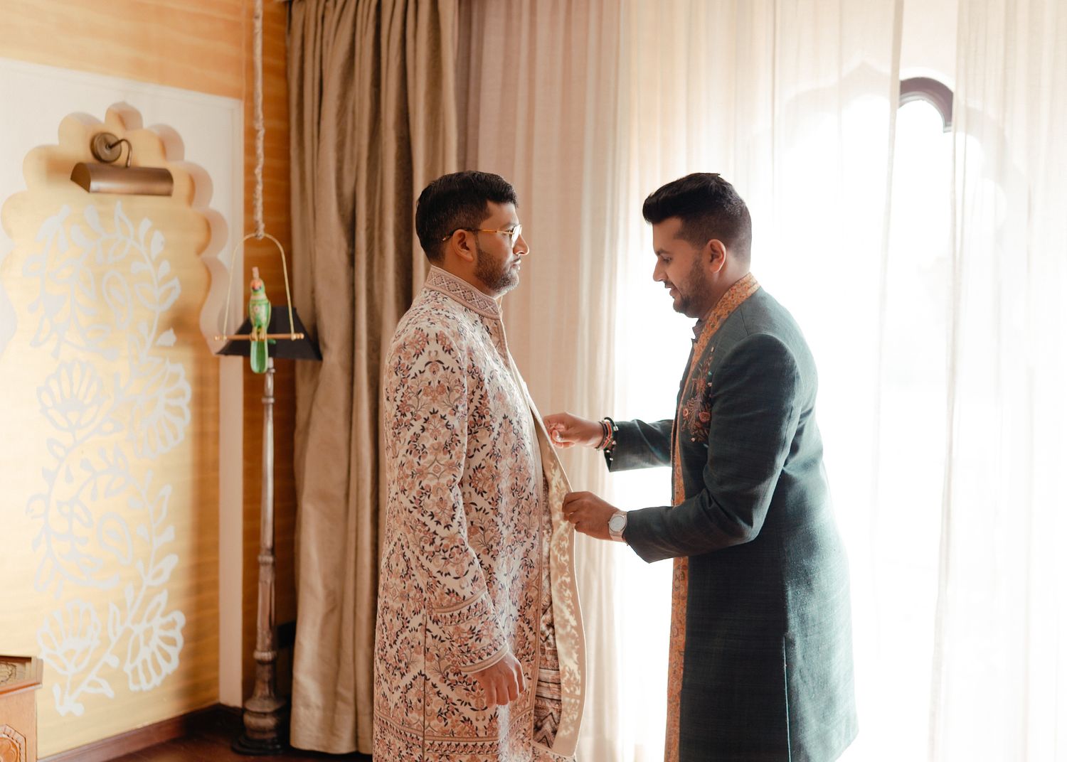Two people in formal attire stand in a sunlit room as one helps adjust the other's white embroidered sherwani.