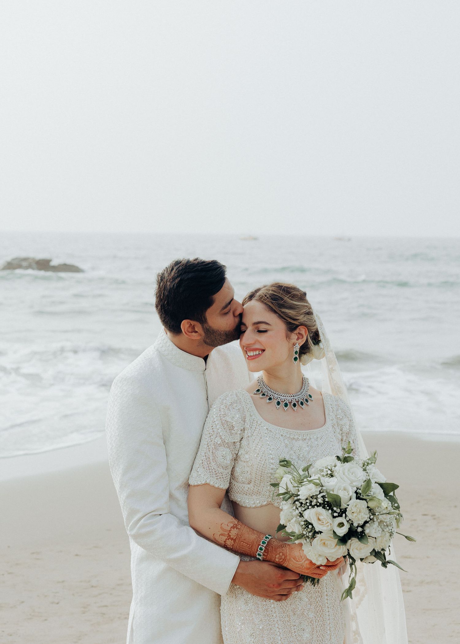 Wedding couple embracing on beach with white floral bouquet and ocean waves behind them.