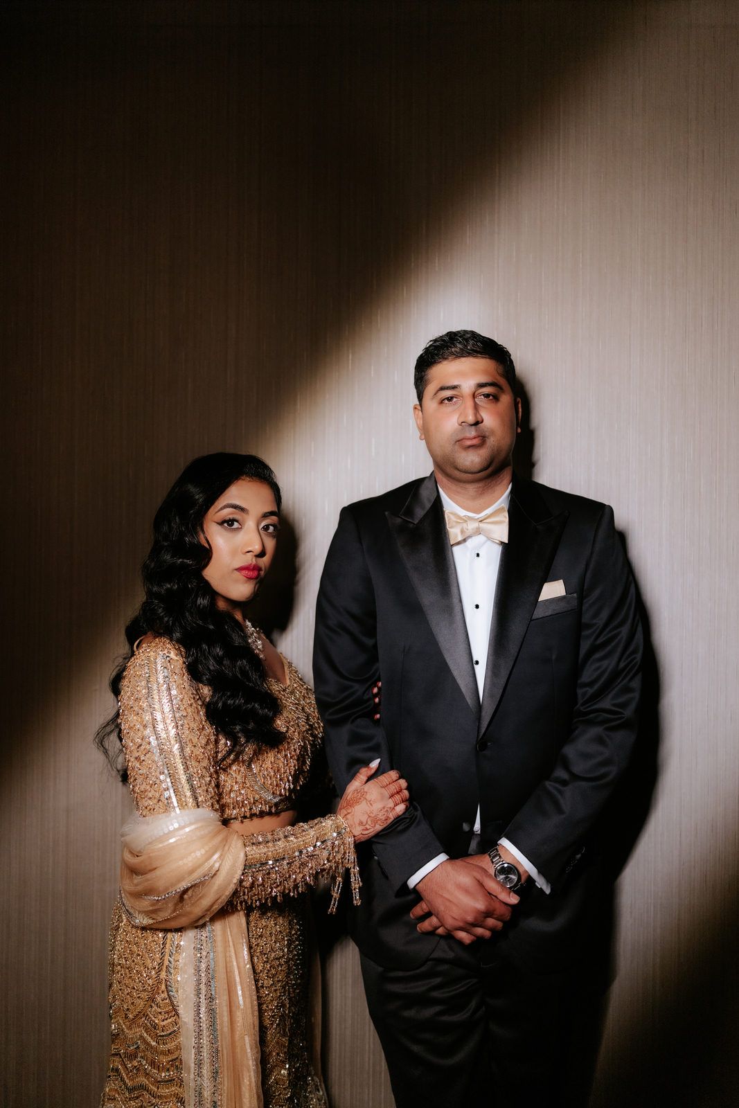 Bride in gold embroidered lehenga and groom in black tuxedo posing together against a neutral textured wall.