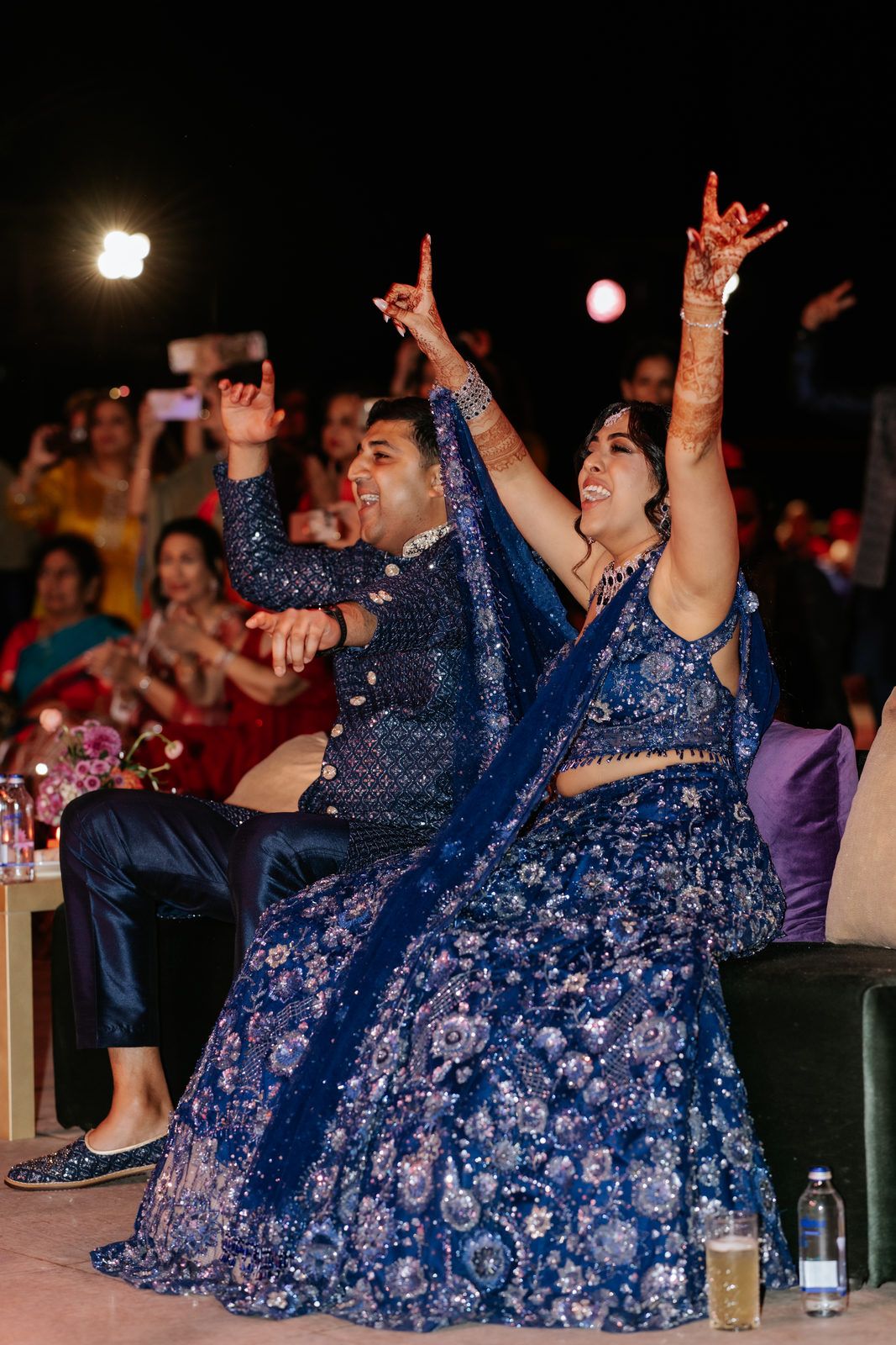 Couple in blue embroidered Indian wedding attire joyfully dancing with hands raised at a festive celebration.
