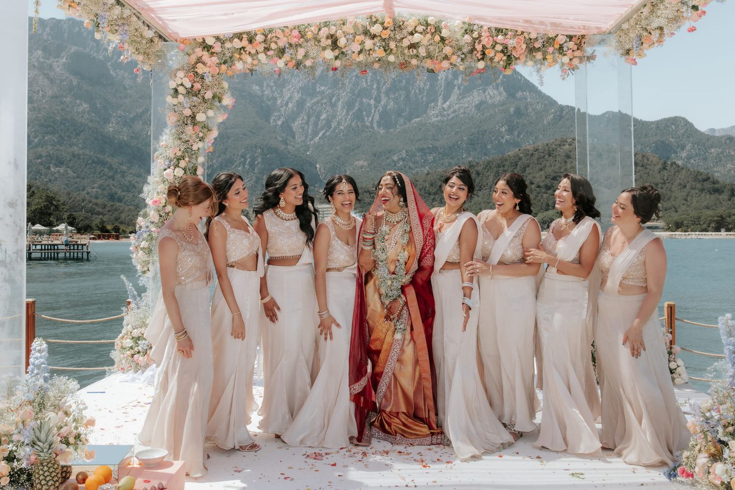 Bride in red saree surrounded by bridesmaids in white gowns under floral mandap with mountain lake backdrop.