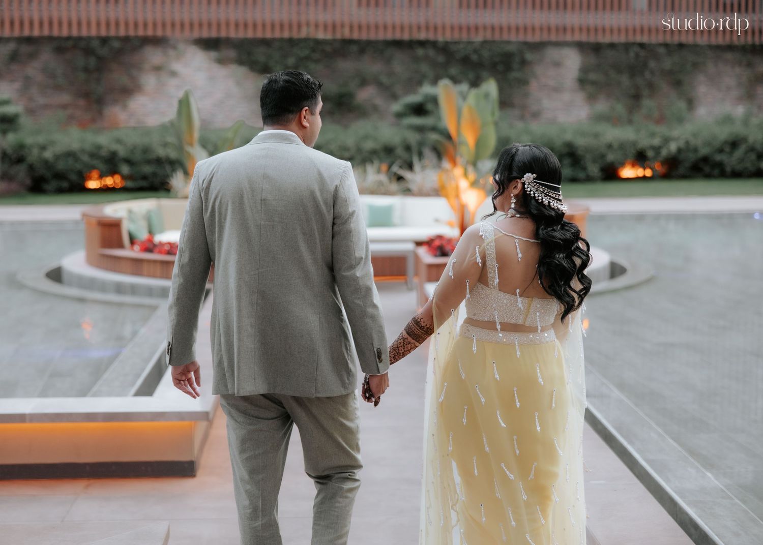 A couple in wedding attire holds hands walking toward an outdoor fire feature at their elegant venue.