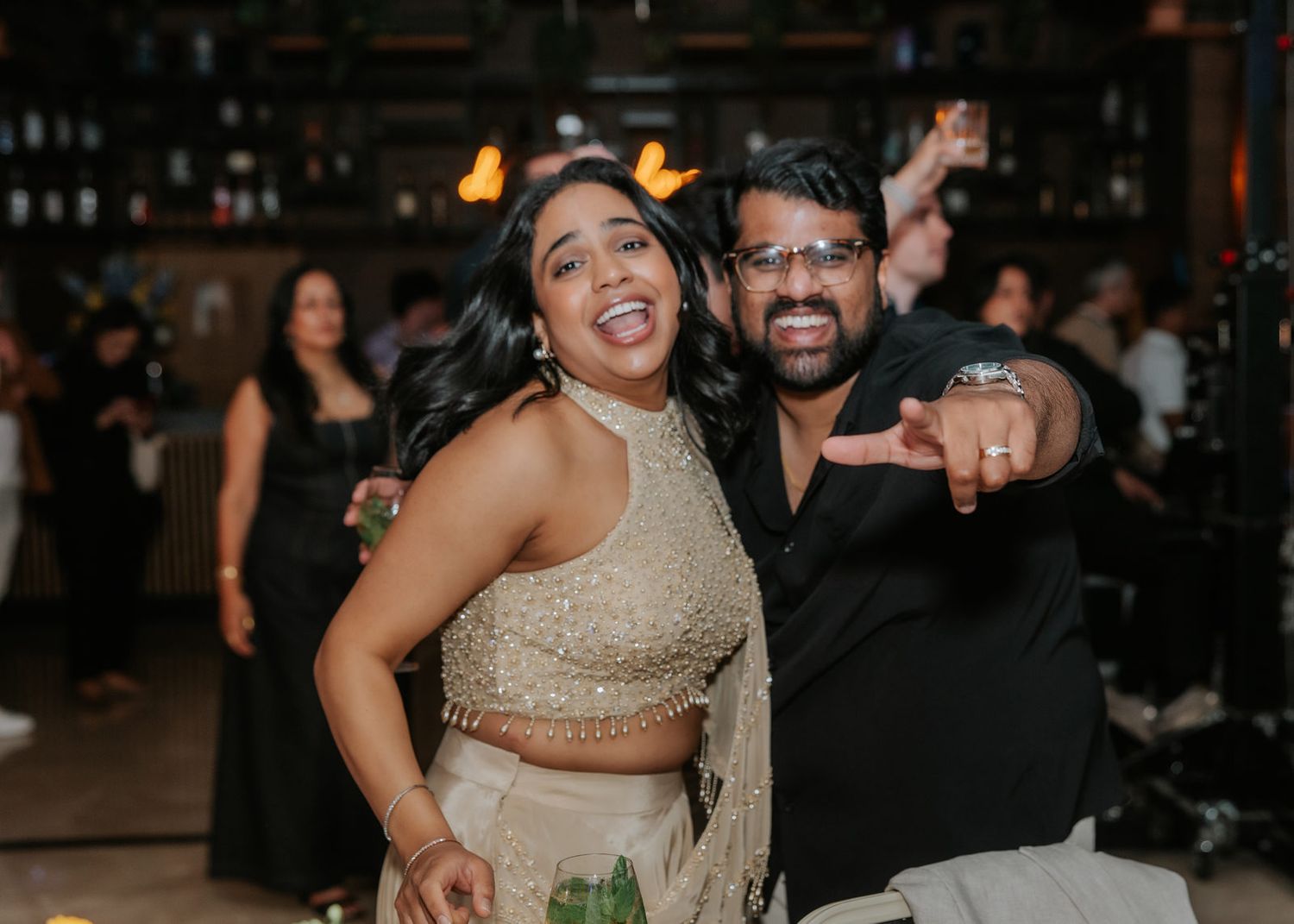 Two people smiling and posing together at an indoor party, woman in gold lehenga and man in black shirt.