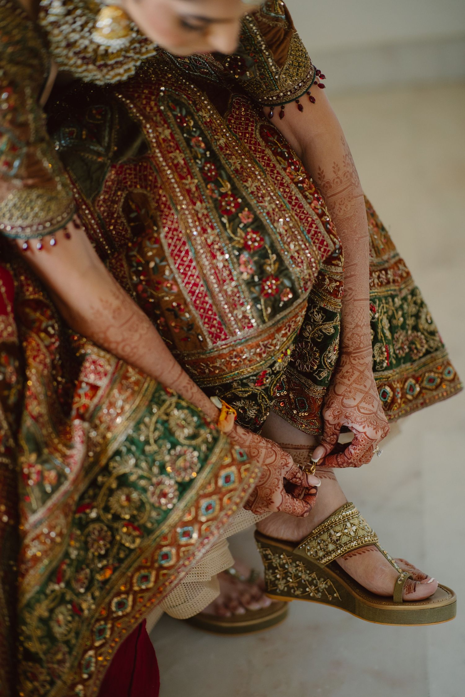Bride in ornate green and red lehenga with mehndi hands fastening gold embellished wedge sandals.