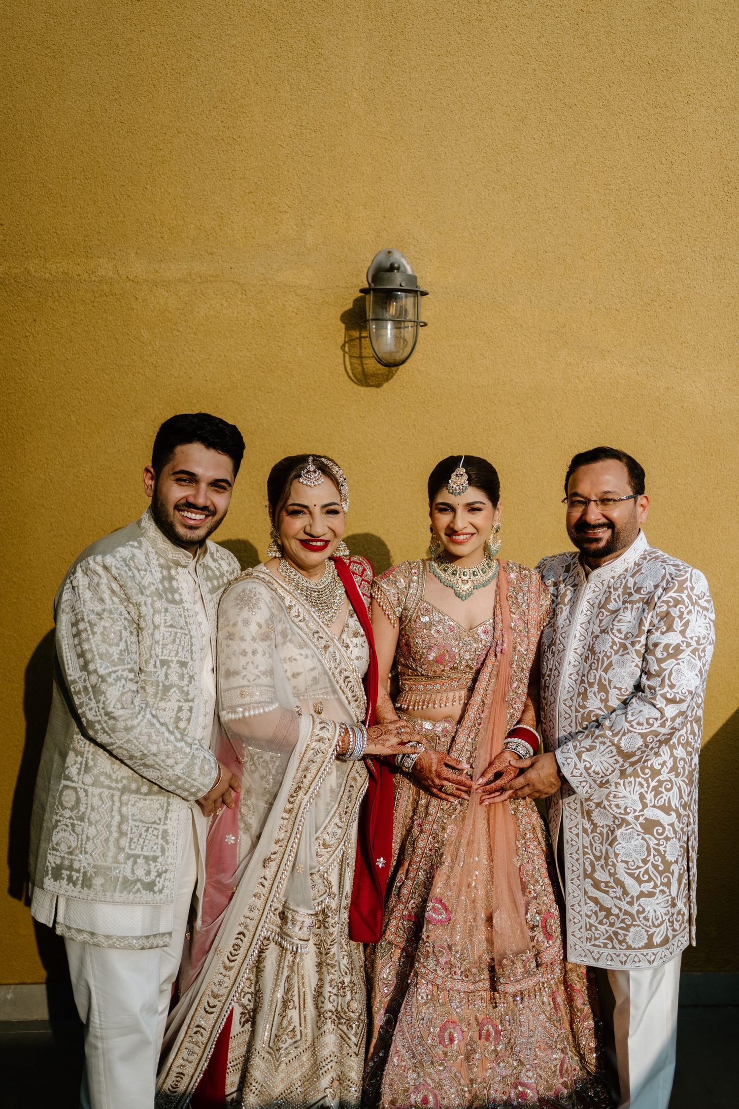 A group wearing elegant white and pink traditional Indian wedding attire poses together against a golden wall.