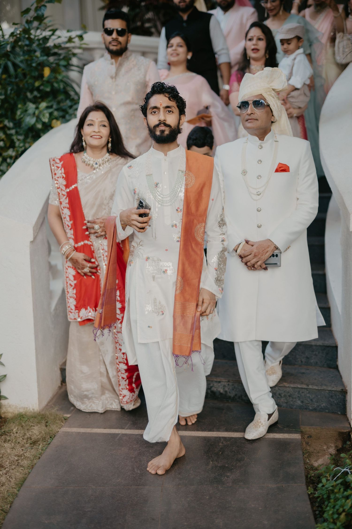 Indian wedding procession with groom in white sherwani, family in traditional attire descending steps.