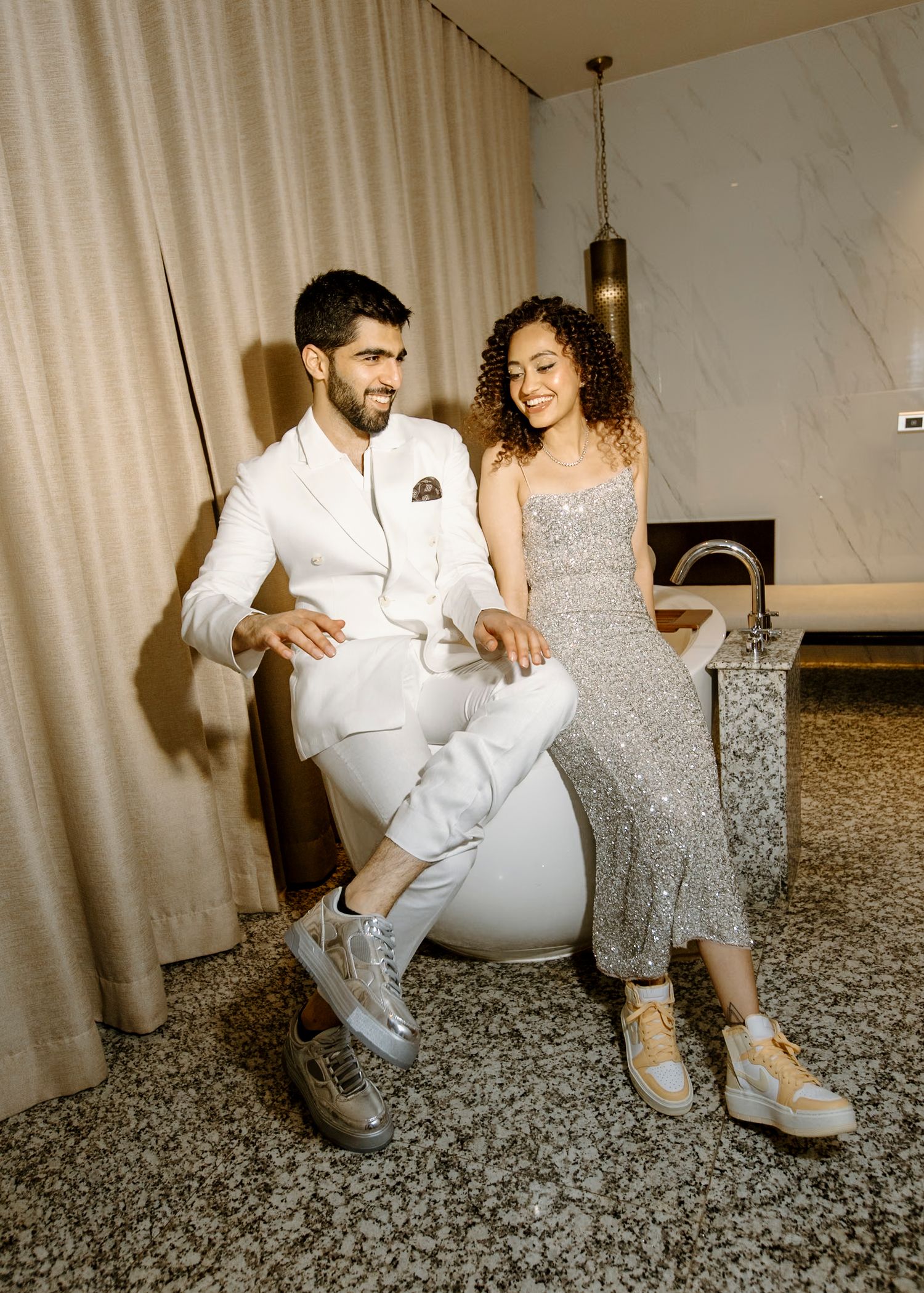 Modern couple's photoshoot featuring white suit and silver dress against marble backdrop.