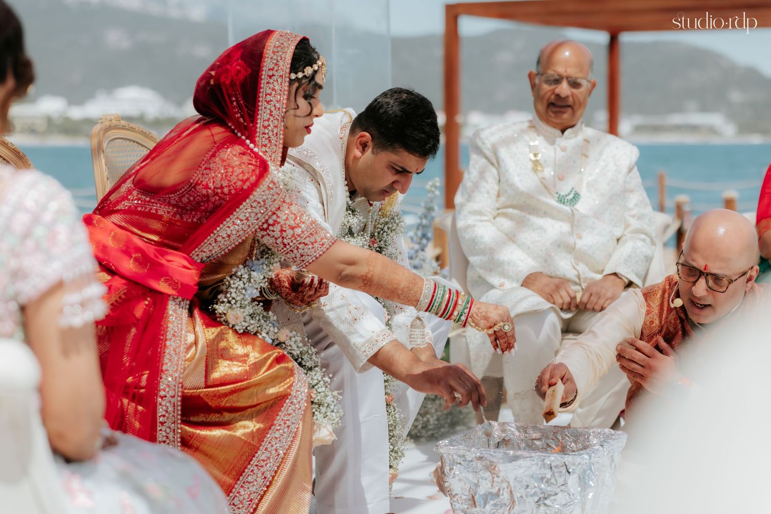 Bride in red saree and groom perform traditional Hindu wedding ceremony outdoors with family seated nearby.