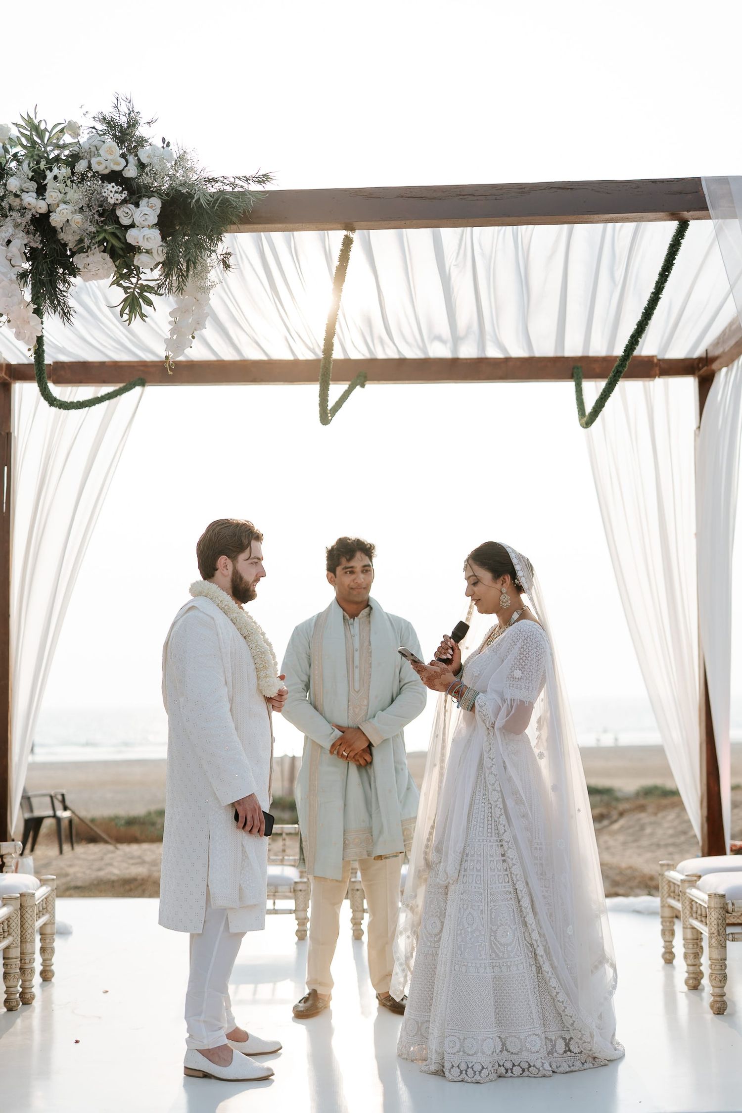 Couple exchanging vows at a beach wedding ceremony under a white draped mandap with floral decor.