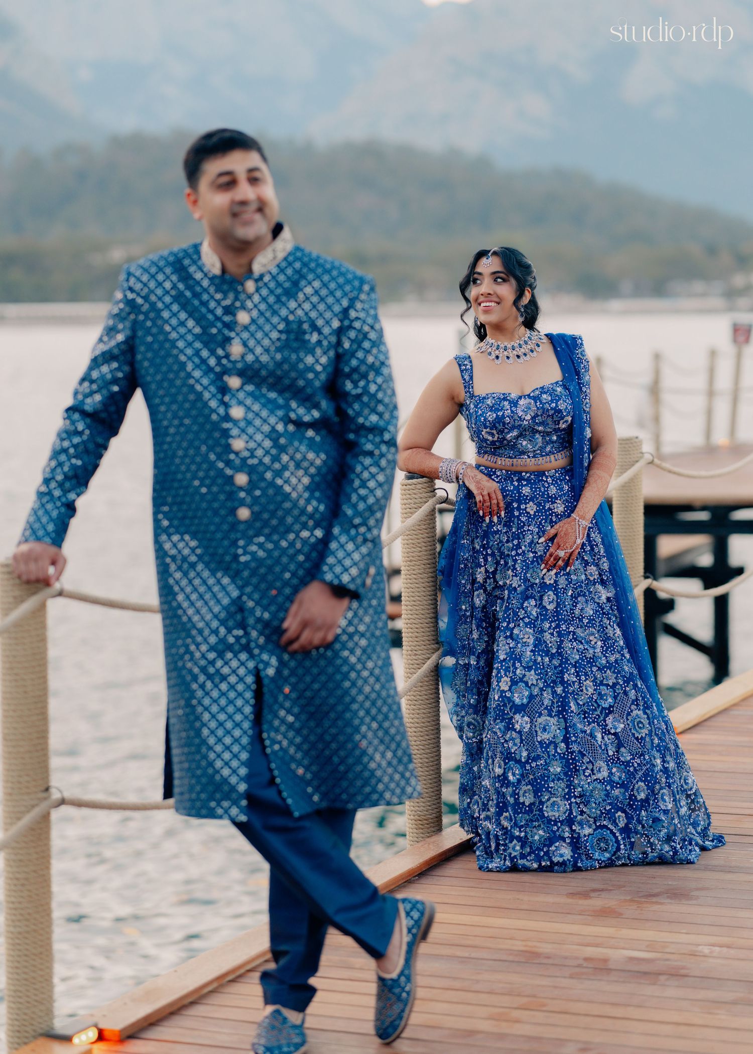 Indian couple in matching blue outfits on a lakeside dock, groom in sherwani and bride in lehenga.