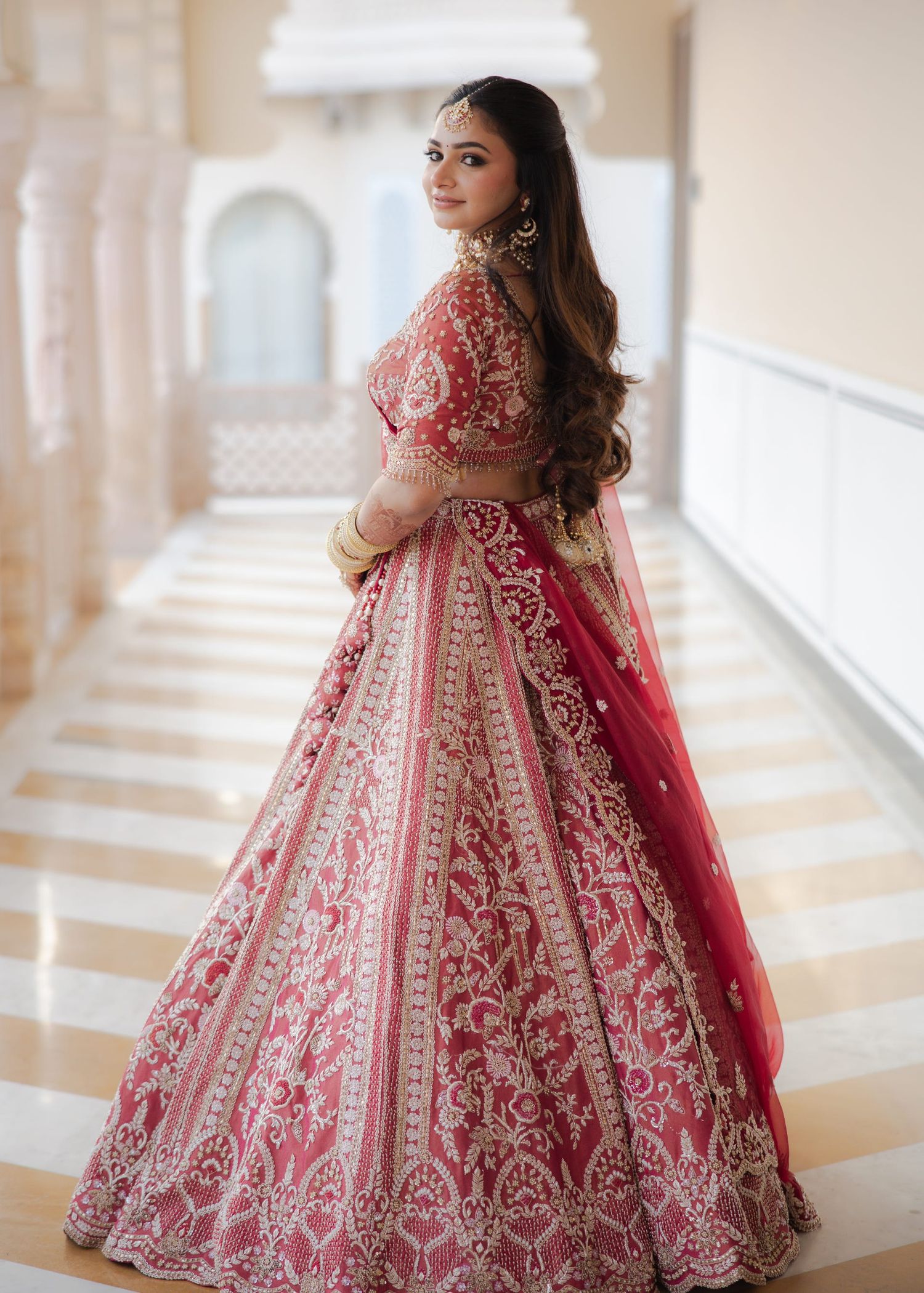 Woman in embroidered red bridal lehenga with gold detailing poses gracefully on marble staircase.