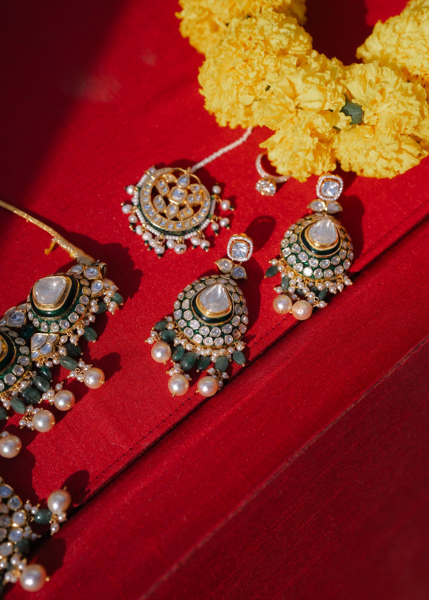 Elegant Indian chandbali earrings with green gems, pearls, and kundan work on red fabric with yellow marigolds.