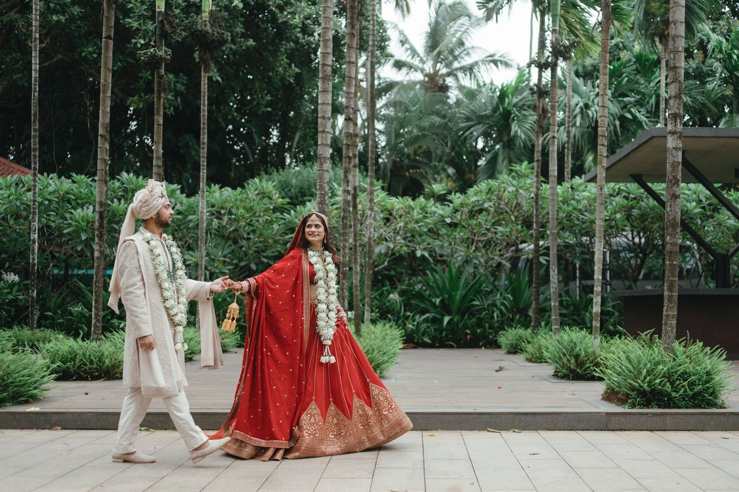Indian bride in red lehenga and groom in white sherwani hold hands walking through lush tropical garden.