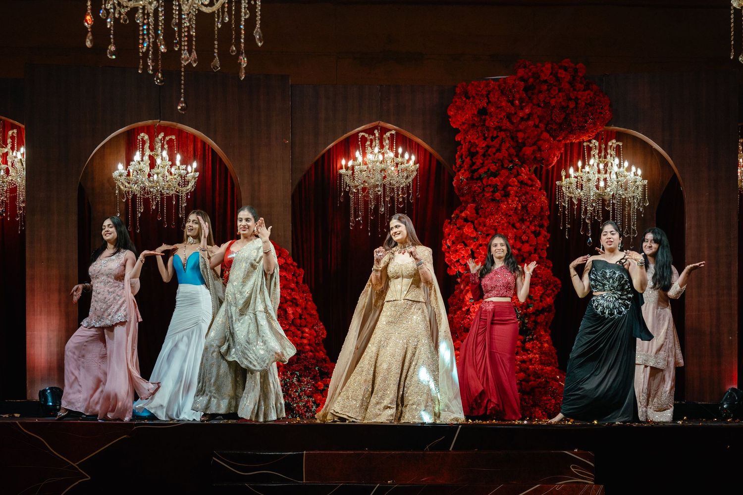 Seven women in elegant gowns perform on a decorated stage with red floral backdrop and chandelier lighting.